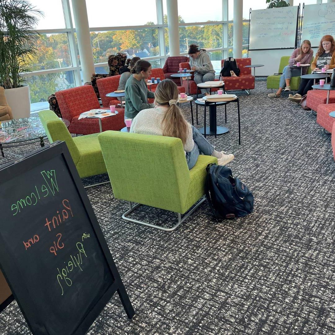 Group of people sitting in chairs, painting, behind sign saying "Paint and Sip Ryleigh"